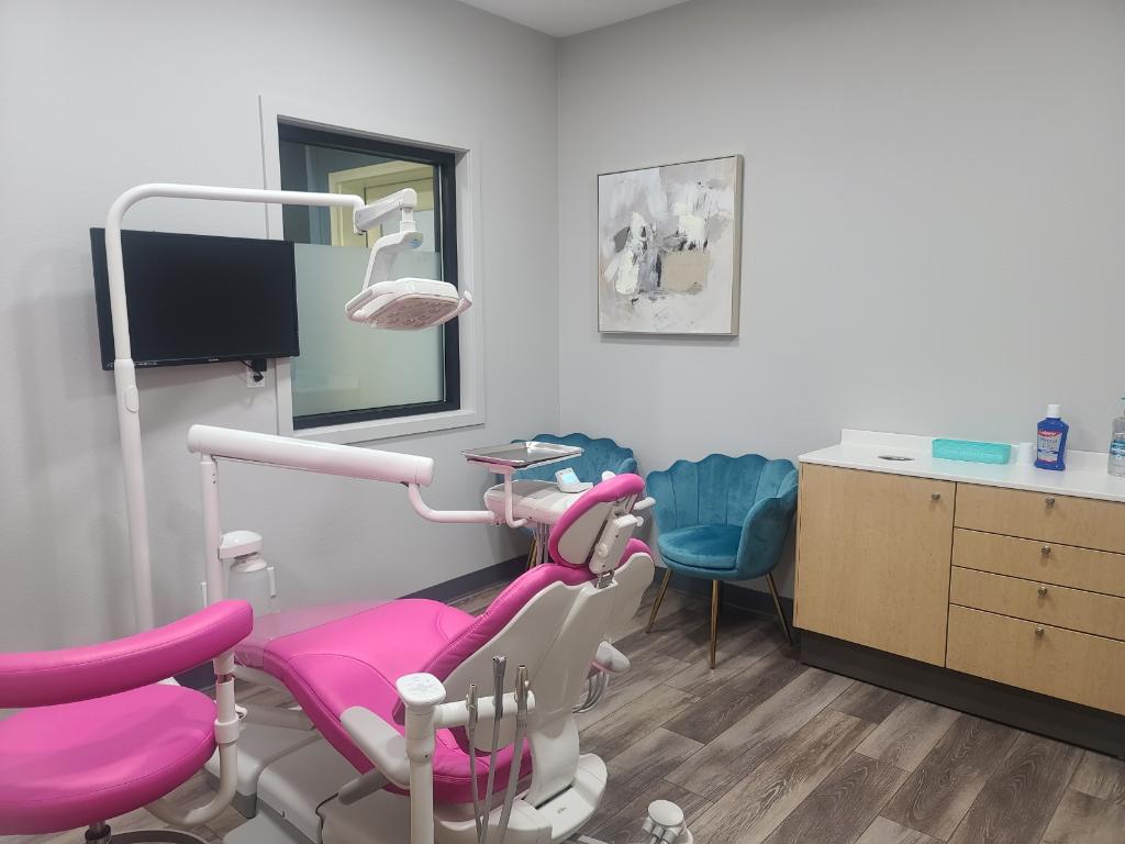 A modern dental examination room featuring a bright pink dental chair, matching stool, overhead light, and a clean, welcoming interior with teal seating and wall art.